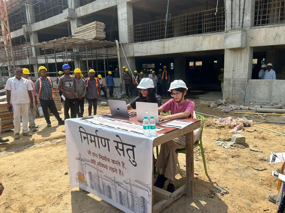Two NirmaanSetu volunteers in hard hats at a registration desk on a construction site, with workers watching