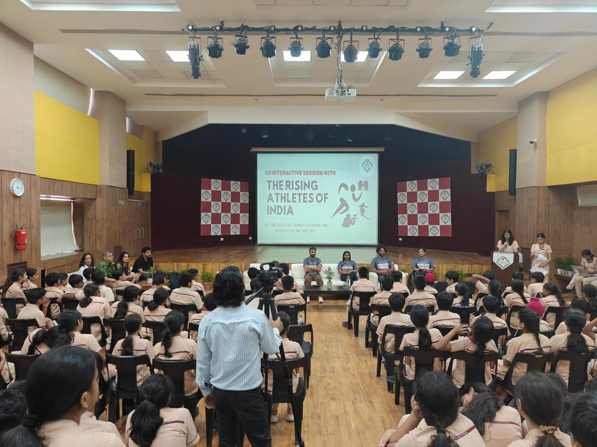 A school hall full of students watching four athletes on a sofa, with a screen reading The Rising Athletes of India